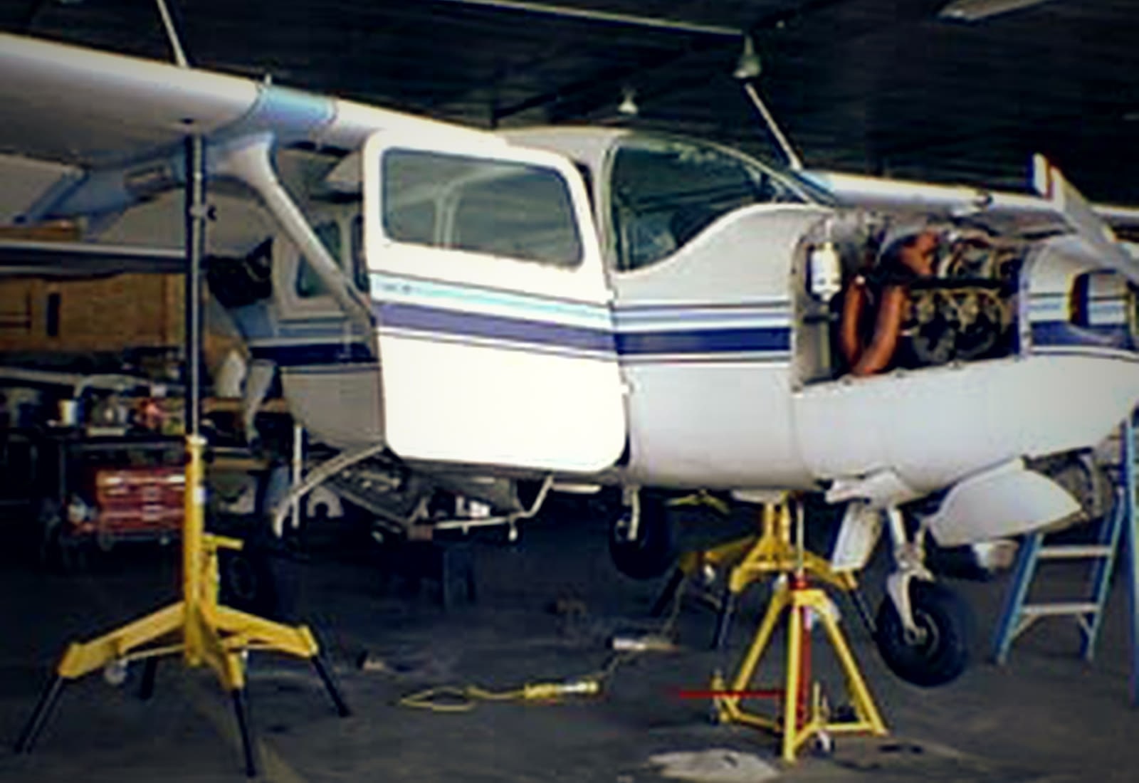 A high-wing Cessna on yellow maintenance jacks in the shop, cowling removed.